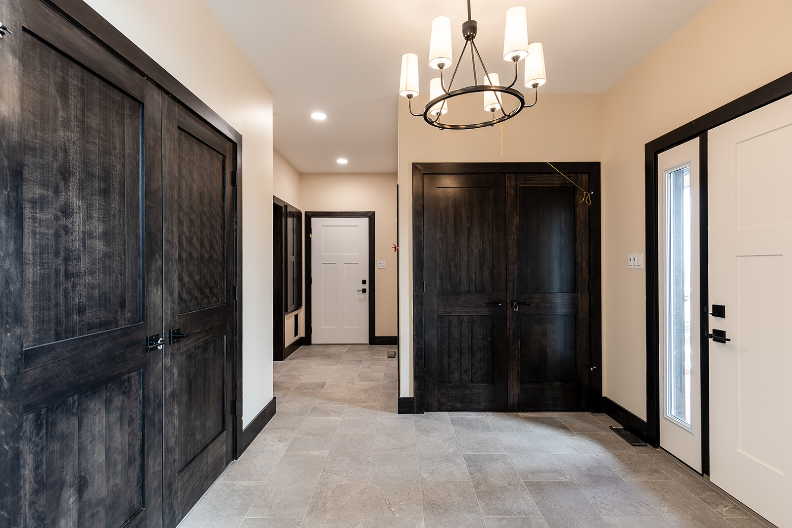 Elegant hallway with light beige walls, dark wooden doors, and gray tiled floor. A modern chandelier hangs from the ceiling, creating a warm ambiance.