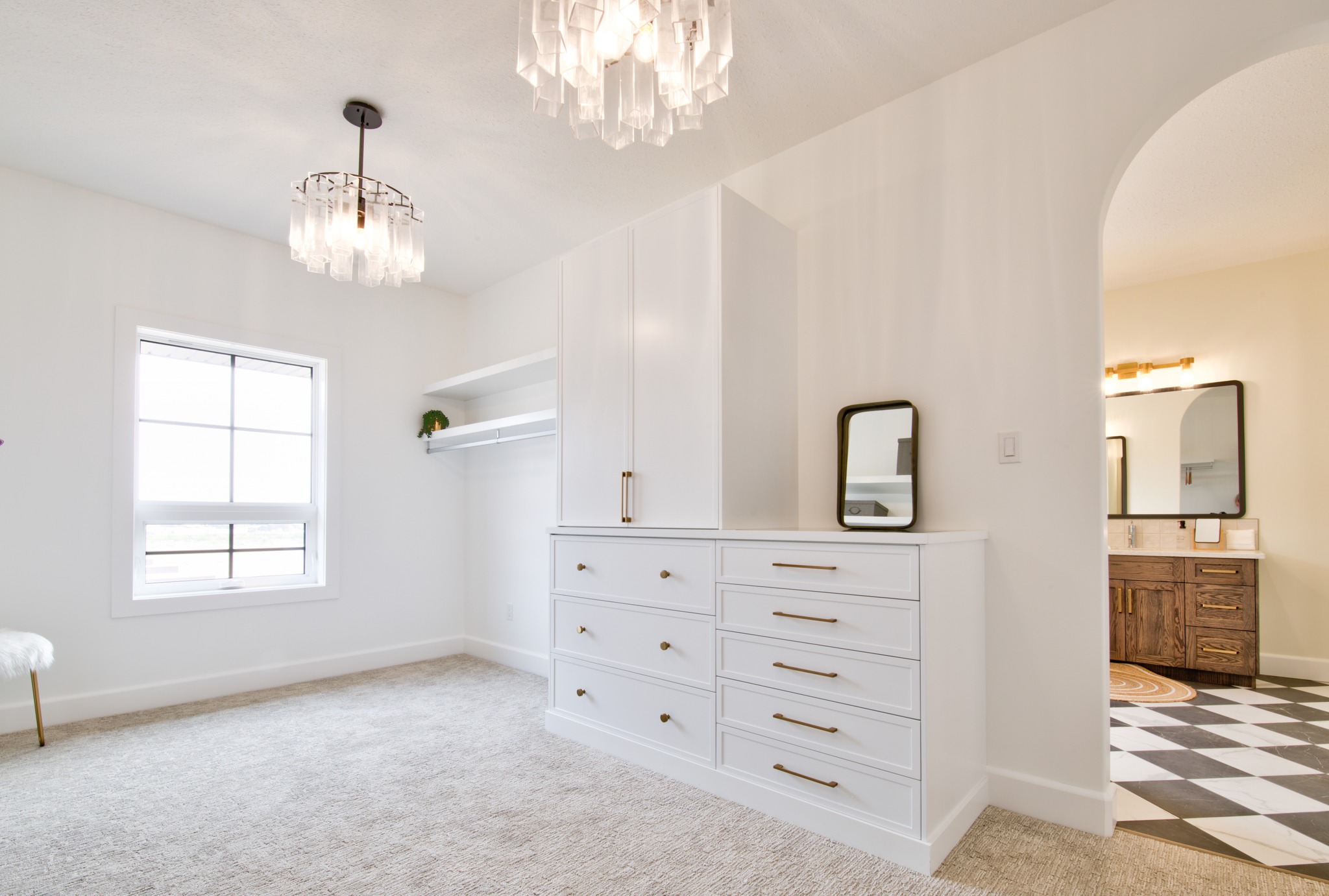 A bright, elegant walk-in closet with white cabinetry, six-drawer dresser, and matching shelves. A modern chandelier hangs overhead. An arched doorway leads to a bathroom with a checkered floor, double vanity, and round mirrors. The atmosphere is clean and sophisticated.