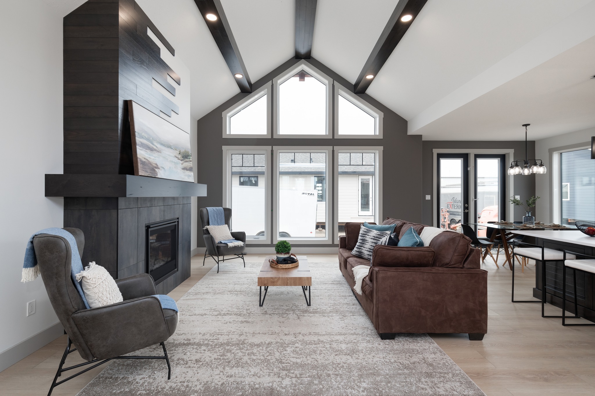 Modern living room with high vaulted ceiling, large windows, and dark wood beams. Features a brown sofa, armchairs, and a sleek fireplace. Bright, airy ambiance.