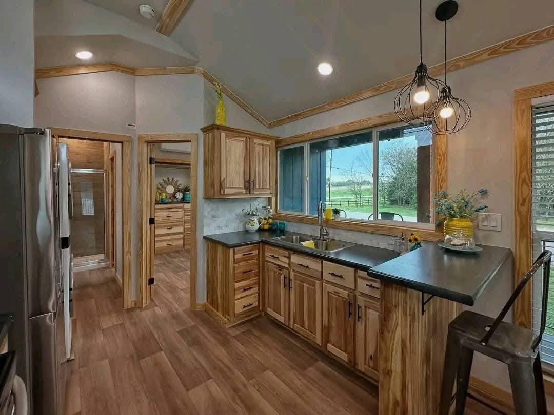 Cozy kitchen with wooden cabinets, dark countertops, and pendant lights. A large window offers a view of a green landscape, creating a warm ambiance.