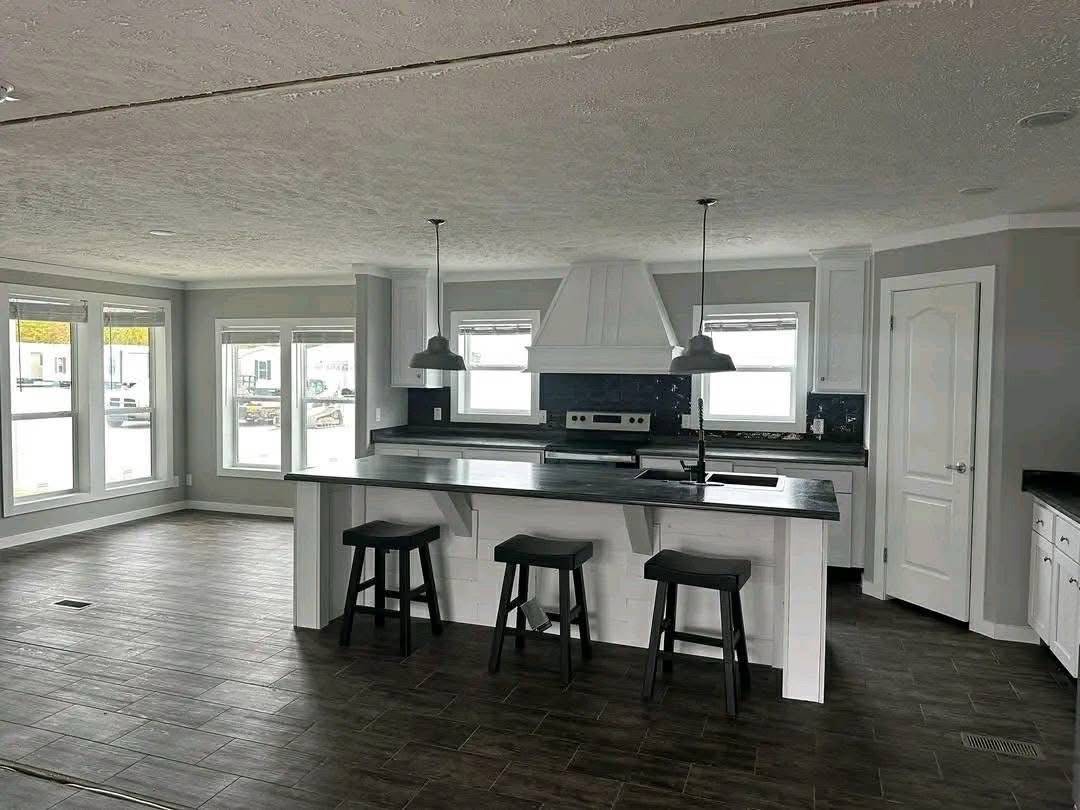 Modern kitchen with white cabinets, dark countertops, and a central island with three stools. Large windows on the left allow ample natural light.