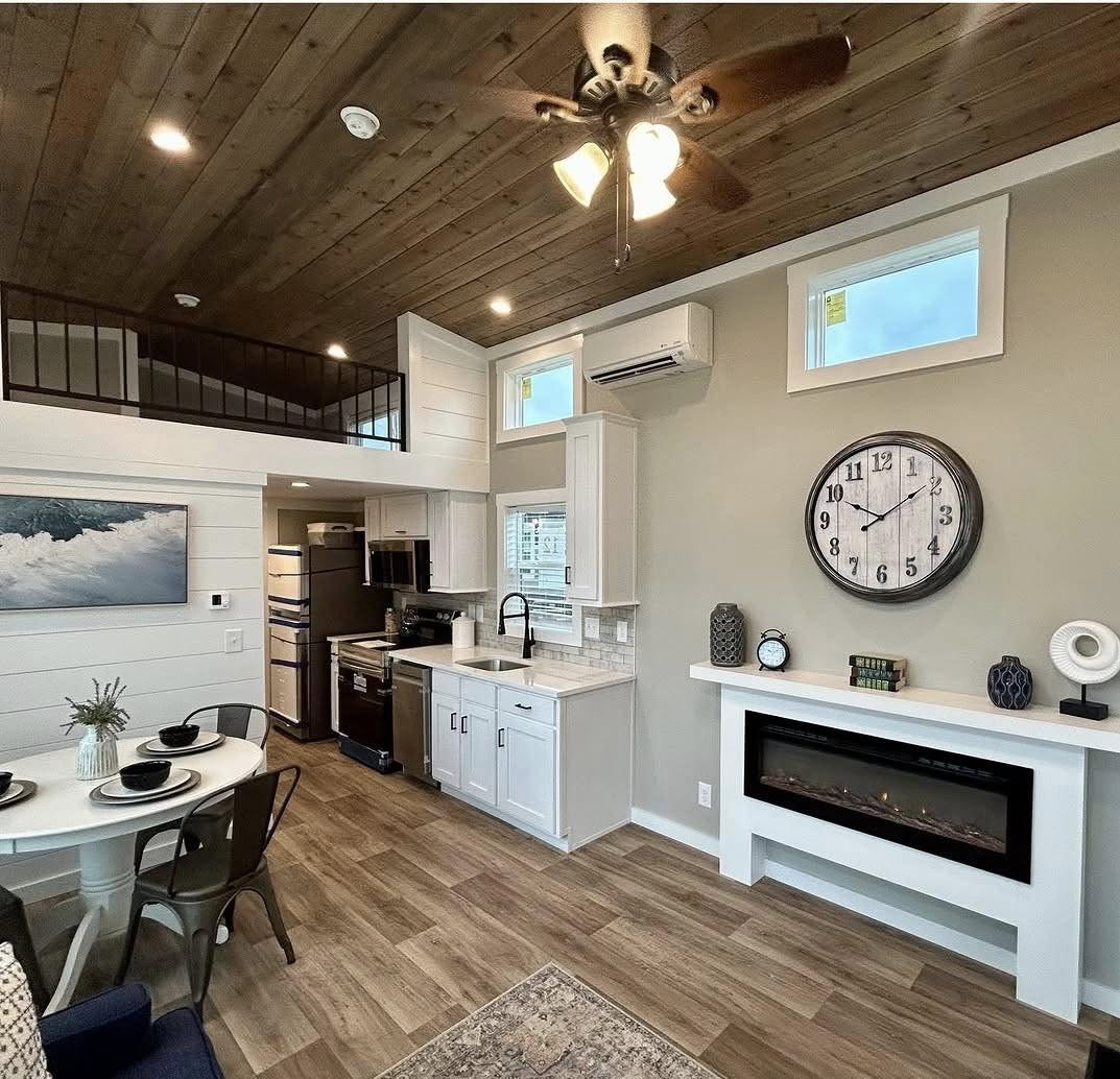 Cozy living space with wooden ceiling and floors, modern kitchen, white cabinets, loft area, large wall clock, and an electric fireplace.