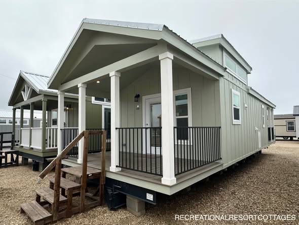 A small, modern tiny house with a green facade and a covered porch sits on a gravel lot. Wooden stairs lead to the entrance, creating a cozy, welcoming feel.