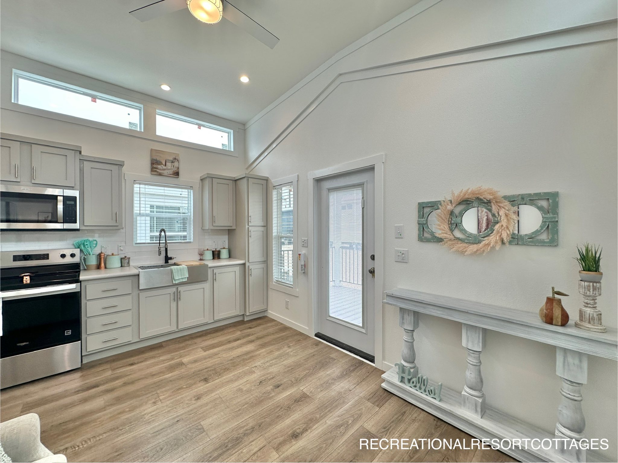 Bright, modern kitchen with light wood flooring, white cabinets, and stainless steel appliances. The room has high ceilings, ample natural light, and a decorative wall shelf with small plants and a mirror.