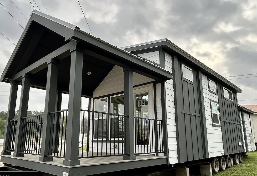 Modern tiny home with gray and white siding on a trailer base. It features a covered porch with black railings under a cloudy sky.