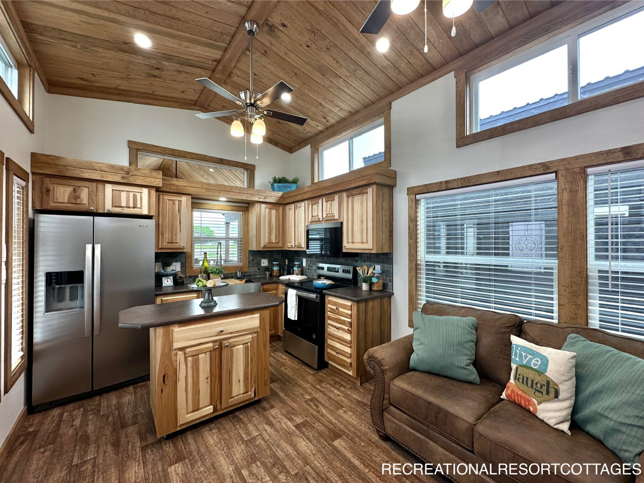 A cozy kitchen with wooden cabinets, stainless steel fridge, and an island. A comfortable couch with colorful pillows sits by large windows. Warm, rustic ambiance.