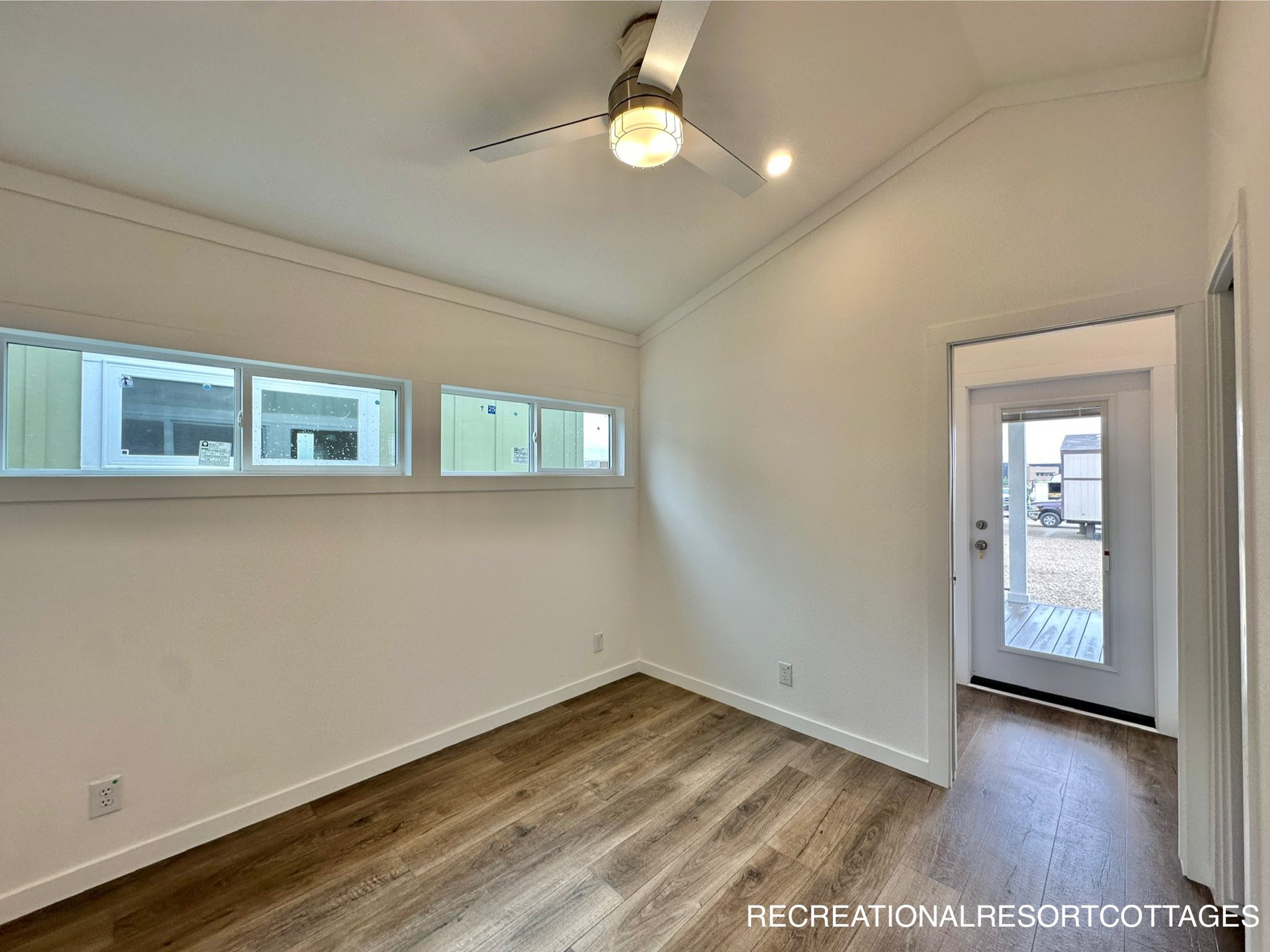 Minimalist room with vaulted ceiling and wood flooring. Horizontal windows on the left, ceiling fan above, and glass door on the right, leading outside.