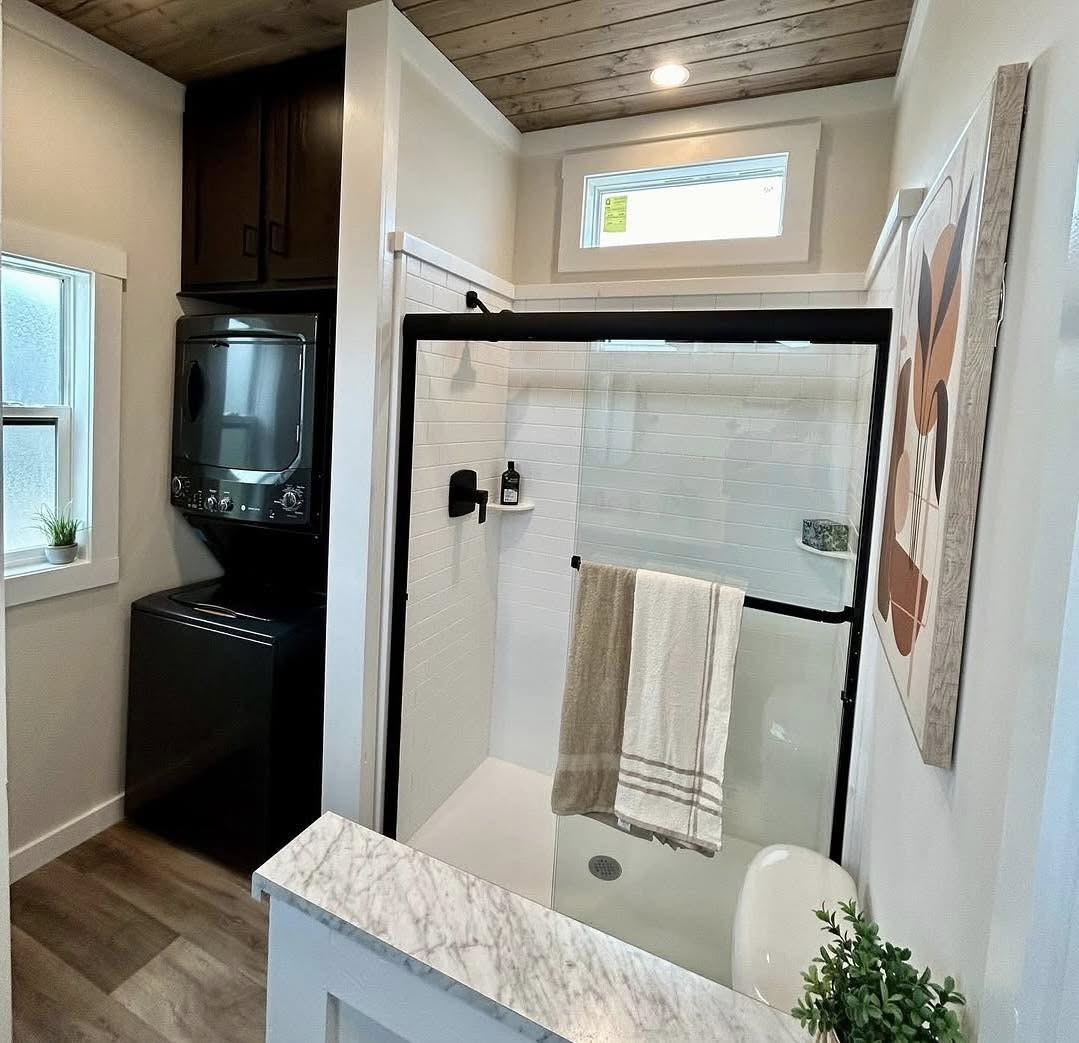 Compact modern bathroom with stacked washer and dryer beside a glass shower. White tiles, wood ceiling, and abstract art create a cozy, clean atmosphere.