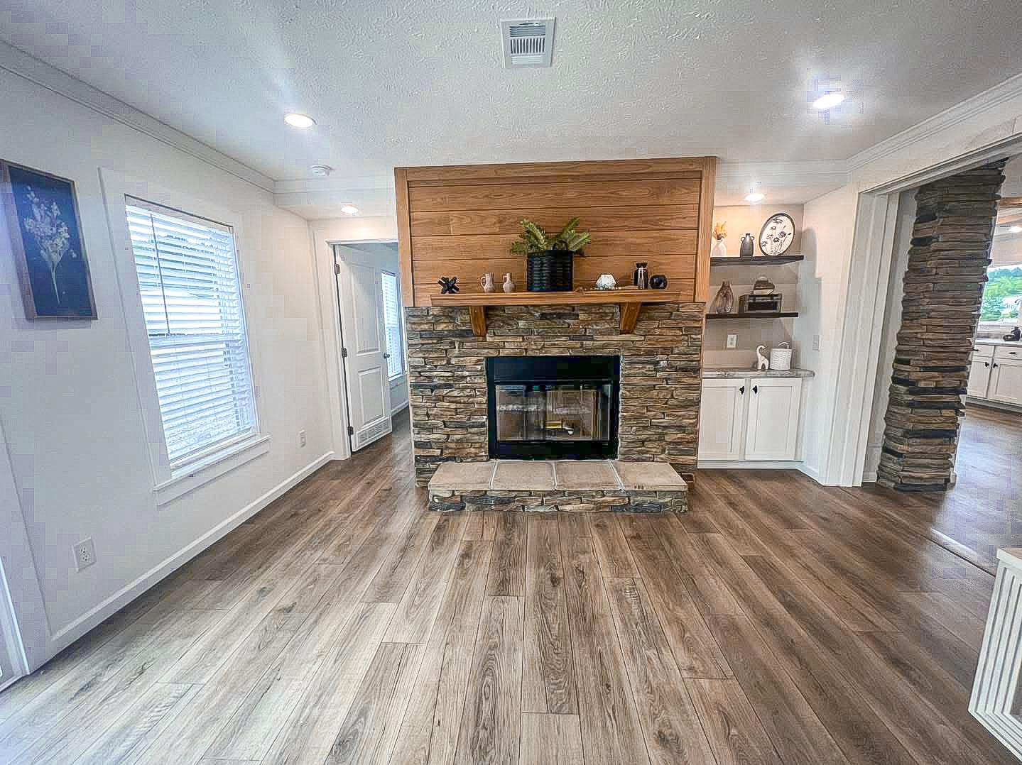 A cozy living room with a stone fireplace and wooden mantel, decorated with plants and ornaments. It features light wood flooring and bright, modern decor.
