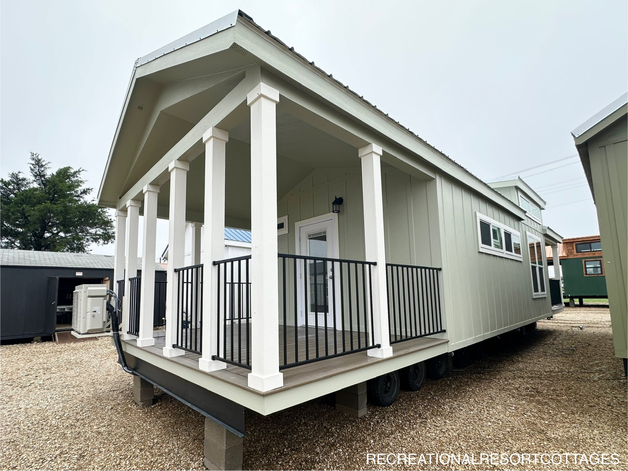 Compact, modern tiny home with a covered porch, white pillars, and black railing. Situated on gravel with overcast sky; a cozy and inviting ambiance.