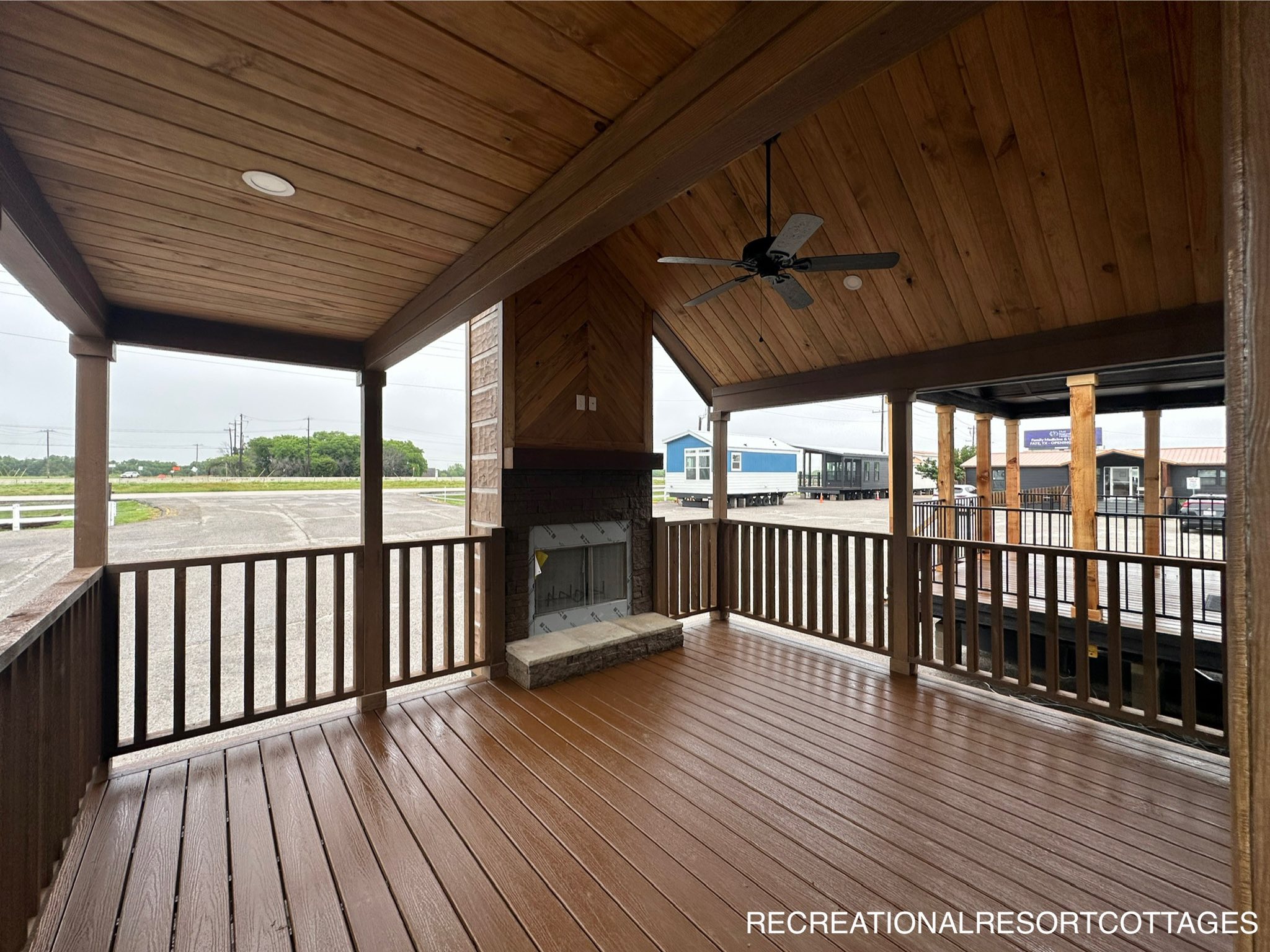 Covered wooden patio with a sloped ceiling, large fan, and a fireplace. Overlooks an empty lot with greenery and parked mobile homes. Calm atmosphere.