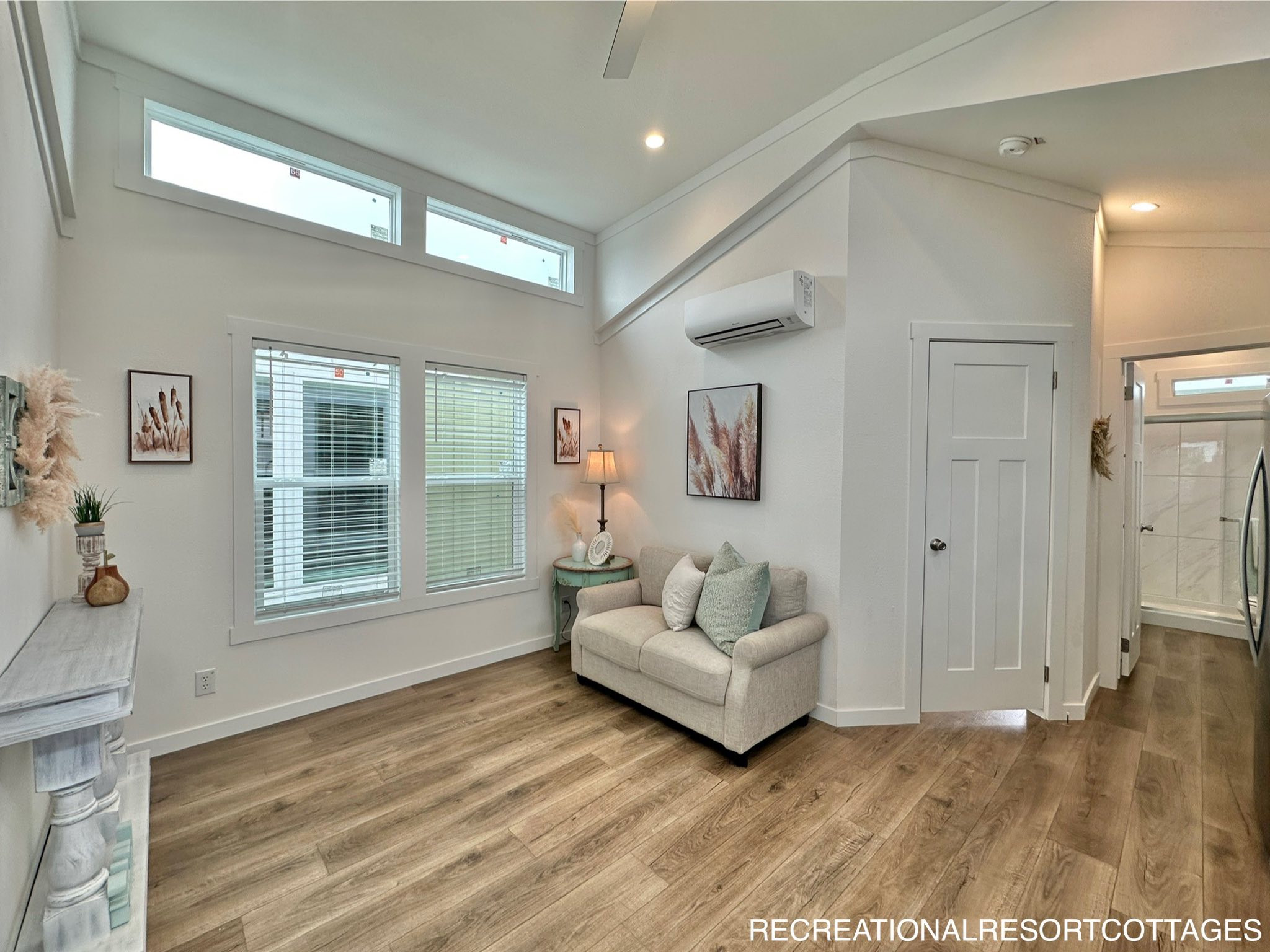 Bright living room with high ceilings, featuring a beige couch with green pillows, a small lamp on a side table, and wooden flooring. Wall art and decor add a cozy touch.