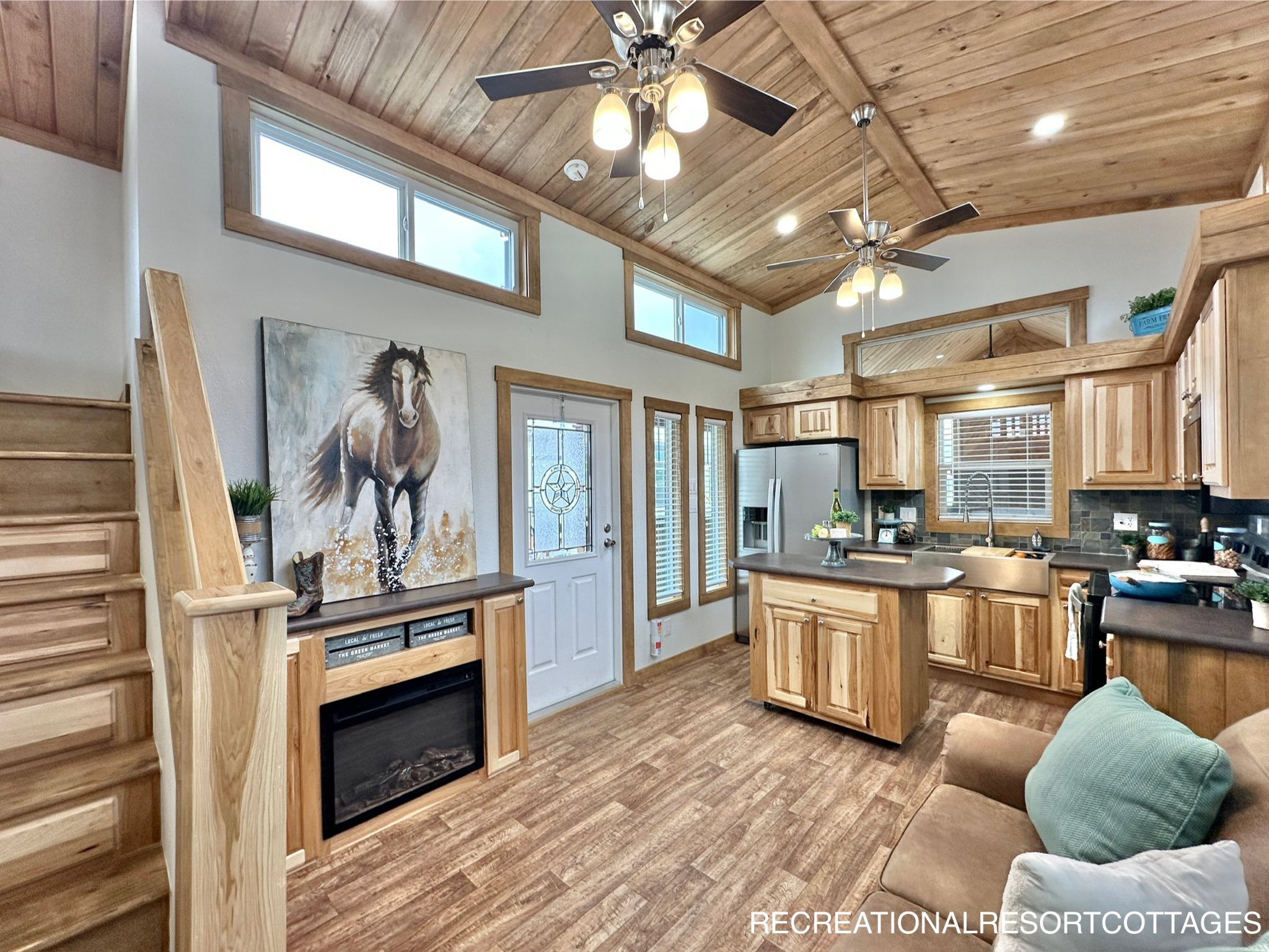 Cozy cabin interior with wood-paneled walls and ceiling, featuring a small kitchen. A horse painting hangs above an electric fireplace by the stairs.
