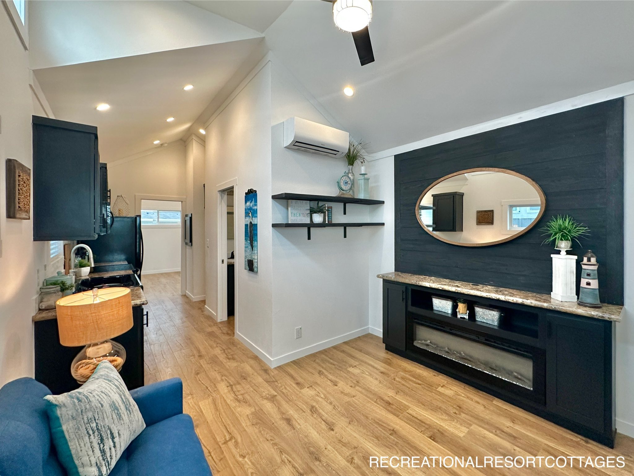 Modern living room with wood flooring and vaulted ceiling. Features a blue sofa, elegant shelves, oval mirror, and stylish console with decor. Bright, cozy ambiance.