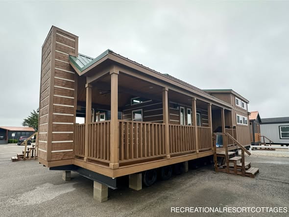 A modern tiny house with a brown wood exterior and a large covered porch on wheels, featuring multiple windows and a chimney. Overcast sky in the background.