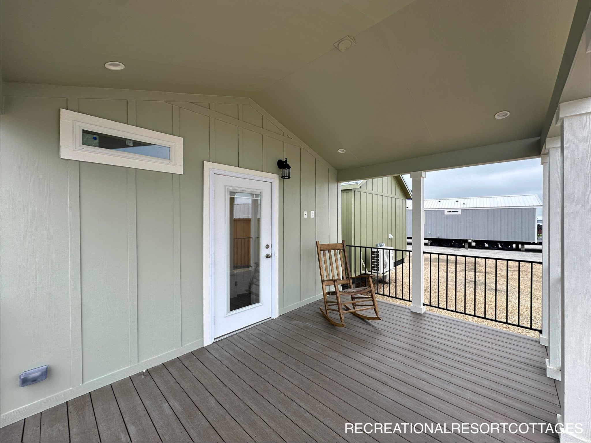 Covered porch with light green siding and a white door, featuring a wooden rocking chair. Open view of neighboring structures under a cloudy sky.