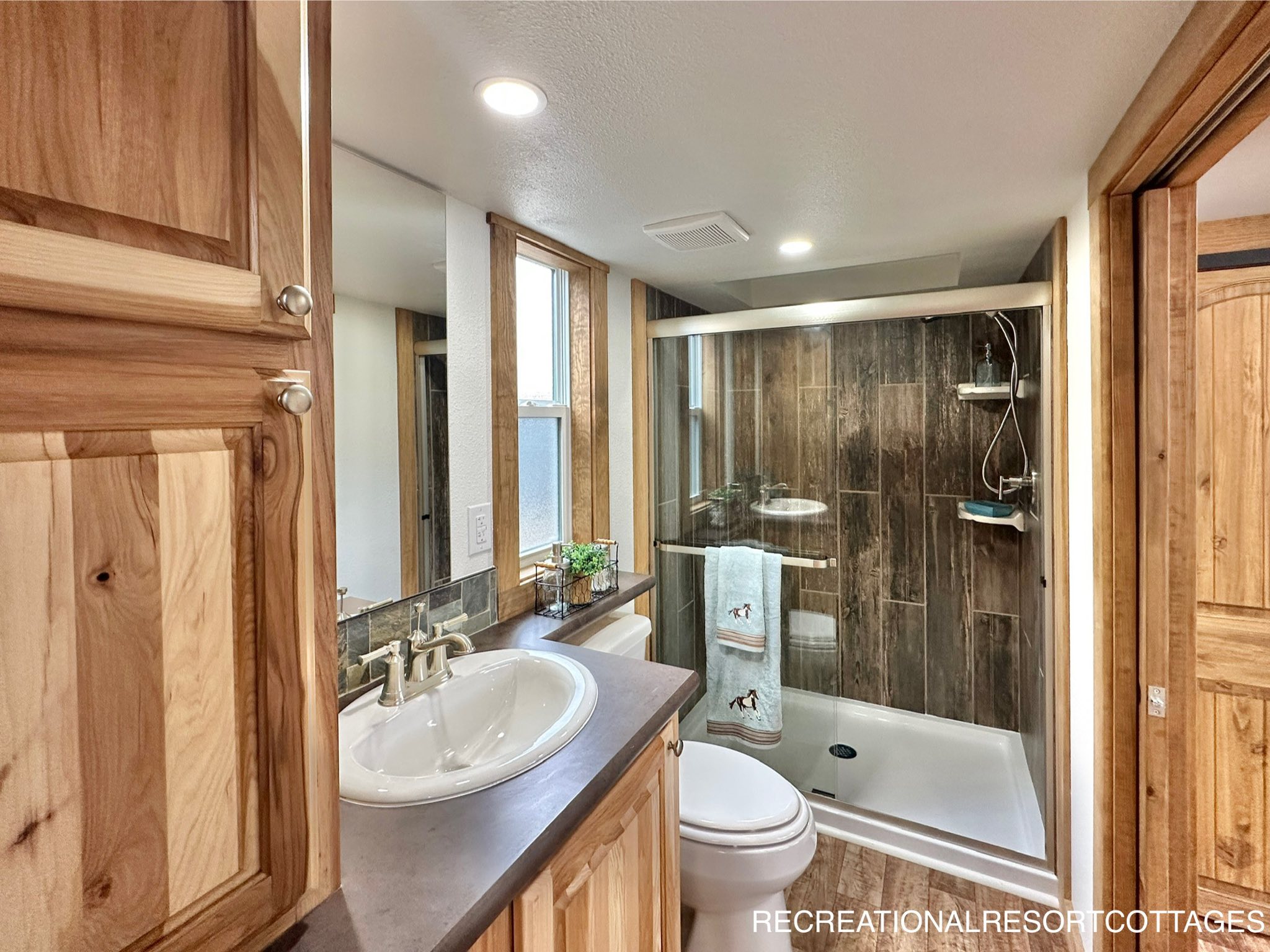 Compact bathroom with wood cabinets, a vessel sink, and a glass shower featuring dark wood tiles. A window adds natural light. Cozy and rustic ambiance.
