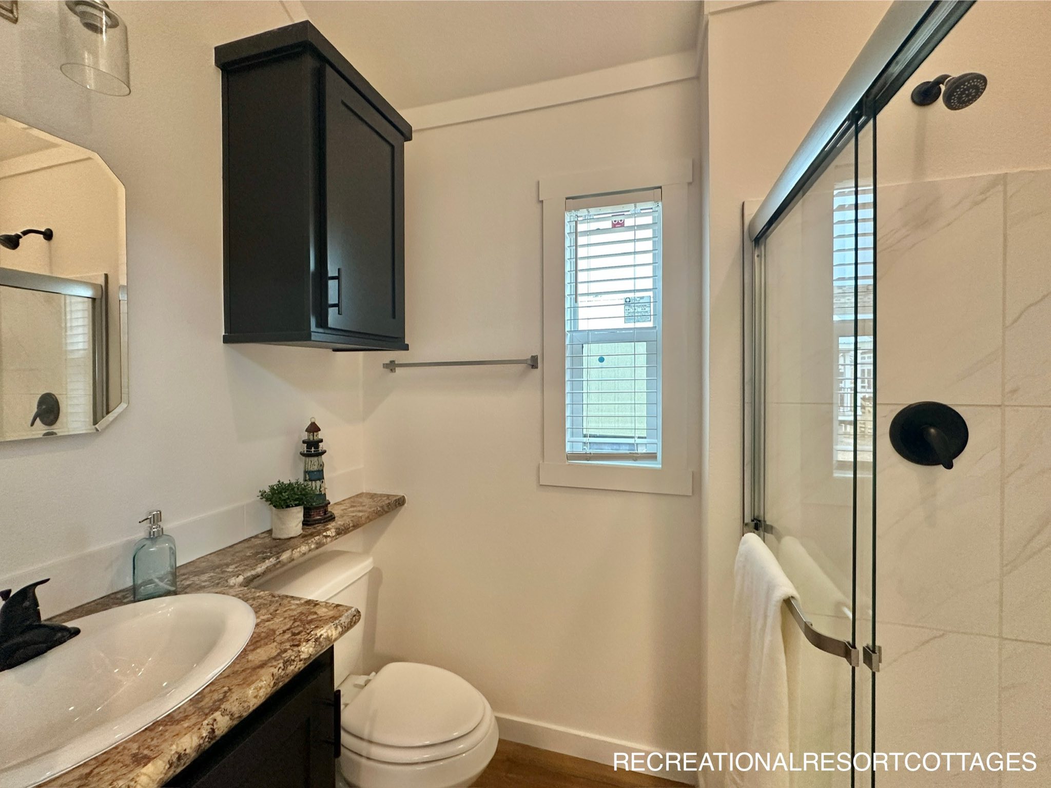 Compact bathroom with white walls, featuring a brown granite countertop with a sink, black upper cabinet, window with blinds, toilet, and glass shower.