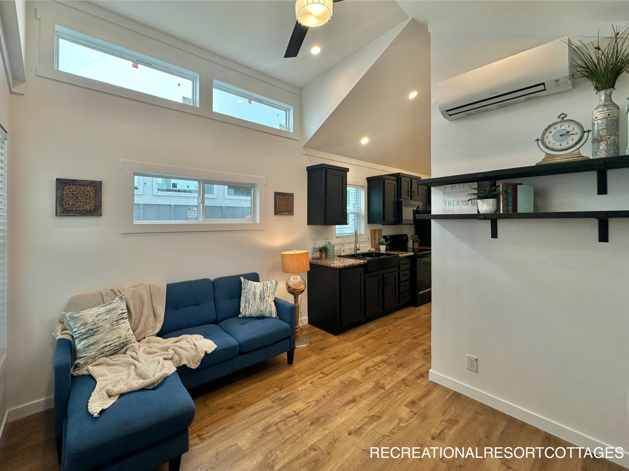 Modern living area in a small cottage. Features a cozy blue sofa with cushions, a throw blanket, wood floor, compact kitchen, and decorative shelves.