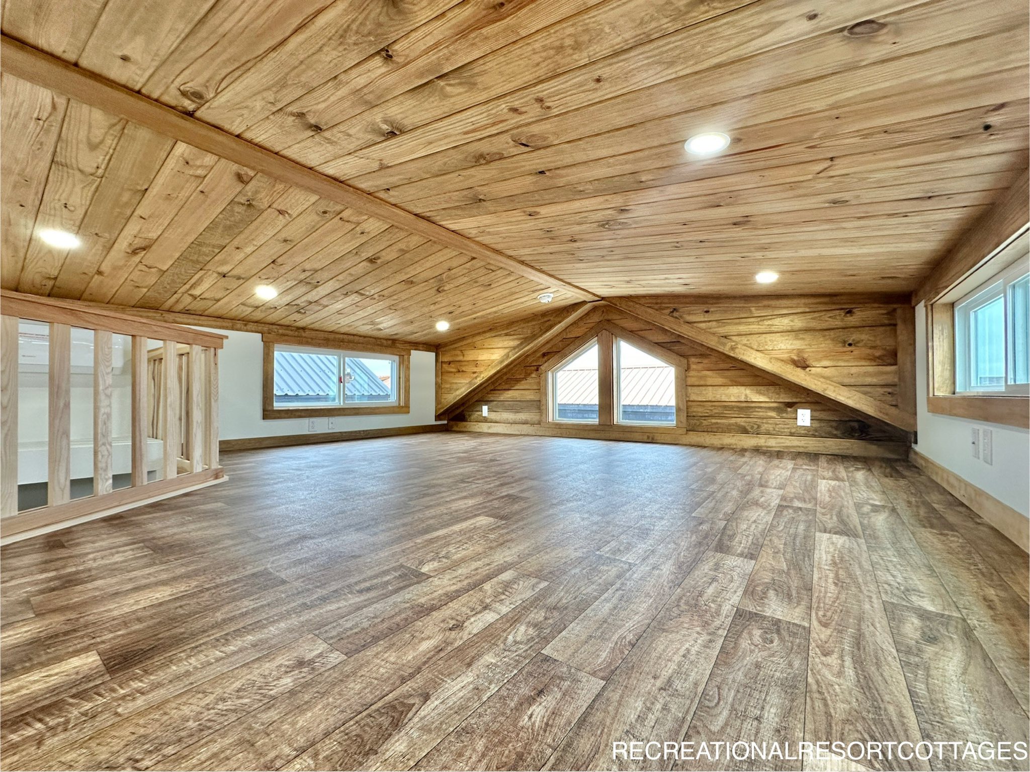 Cozy loft space with wooden ceilings and floors, featuring triangular windows and recessed lighting. Bright and warm atmosphere with rustic charm.