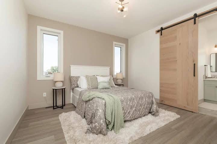 A cozy bedroom with a beige wall features a bed with gray and white bedding, a green throw, and fluffy rug. Two windows flank the bed, and a wooden sliding door leads to a bathroom.