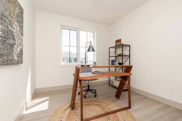 Minimalist home office with a wooden desk centered on a round, woven rug. A metal lamp and book rest on the desk. Bookshelf in the corner, large window letting in natural light.