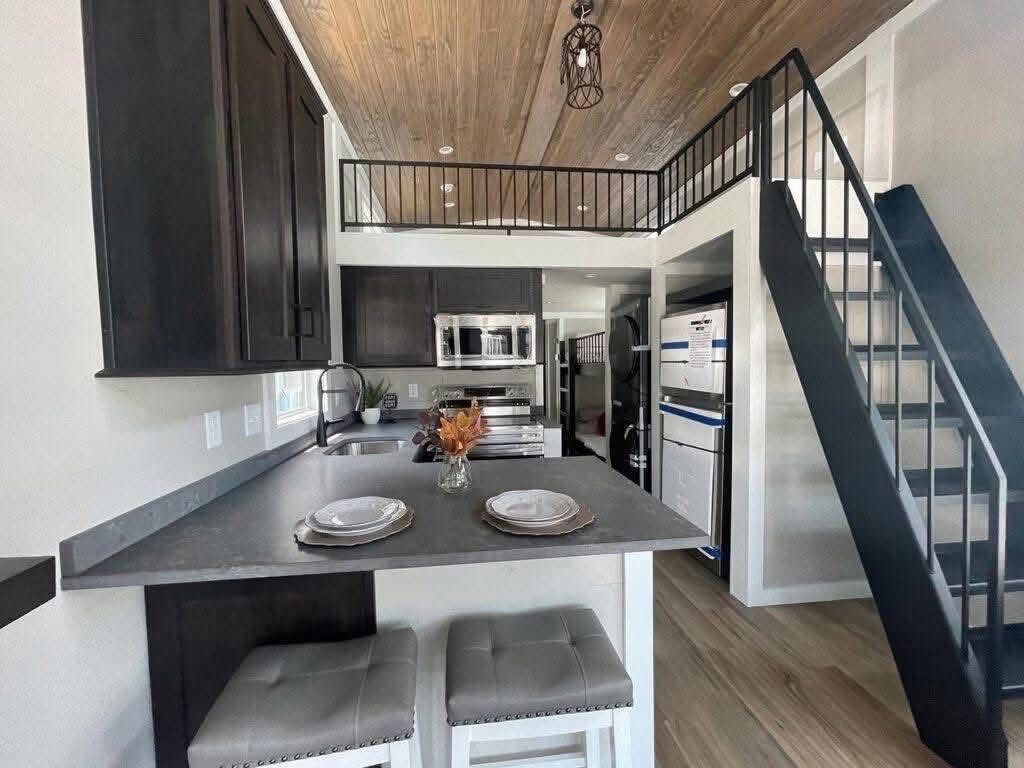 Modern tiny house interior with a loft. The kitchen features dark cabinets, stainless appliances, and a breakfast bar with stools. A staircase leads to a loft.