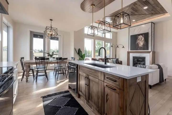Modern kitchen with wood accents, featuring a large island with a white countertop and black faucet. Pendant lights hang above. A dining area with a wooden table and chairs is near large windows, creating a bright and airy atmosphere.