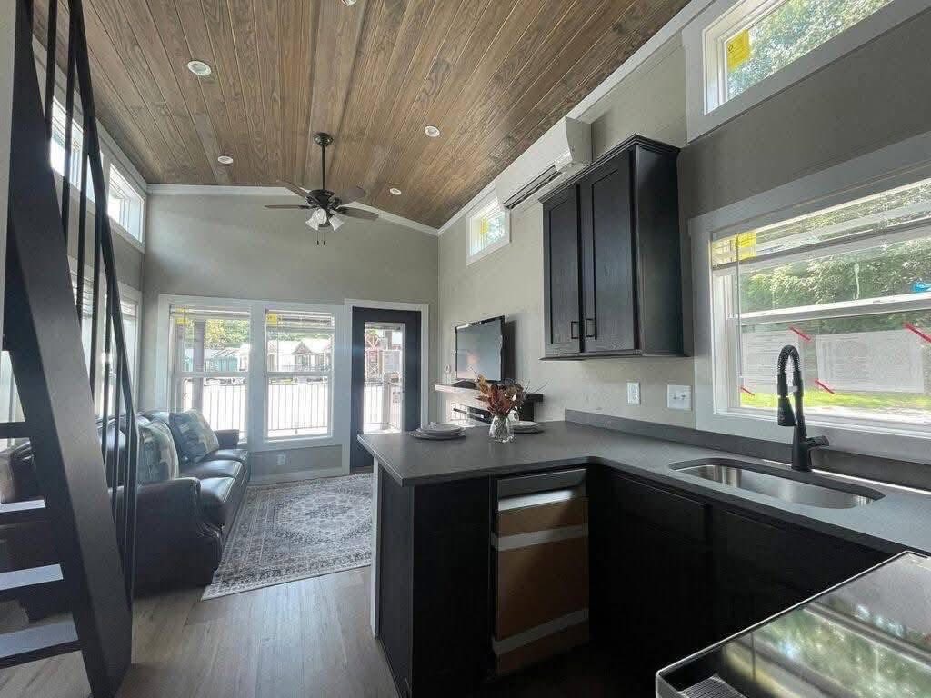 Modern tiny home interior with high ceilings, wood-paneled roof, and large windows. Features a cozy living area, black cabinets, and a compact kitchen. Warm and inviting atmosphere.