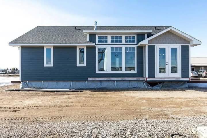A modern, single-story prefab home with blue siding and large windows. It sits on a sandy plot, conveying a sense of simplicity and functionality.