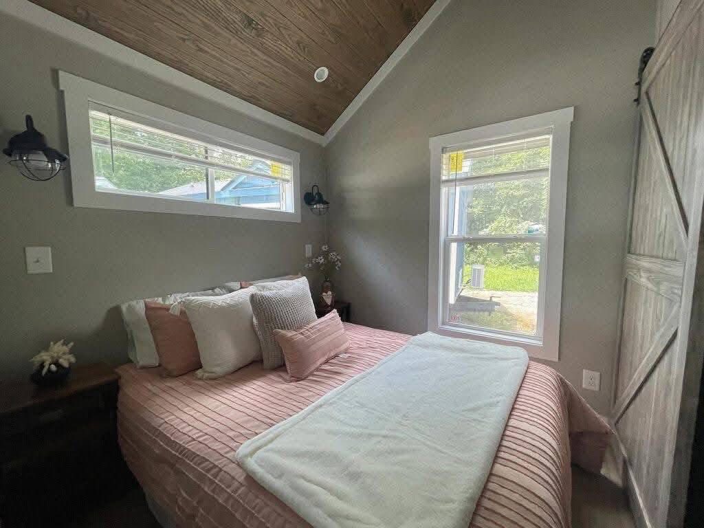 Cozy bedroom with a wooden ceiling, gray walls, and two windows. The bed, adorned with pink and white pillows, adds warmth. Soft, natural light fills the space.