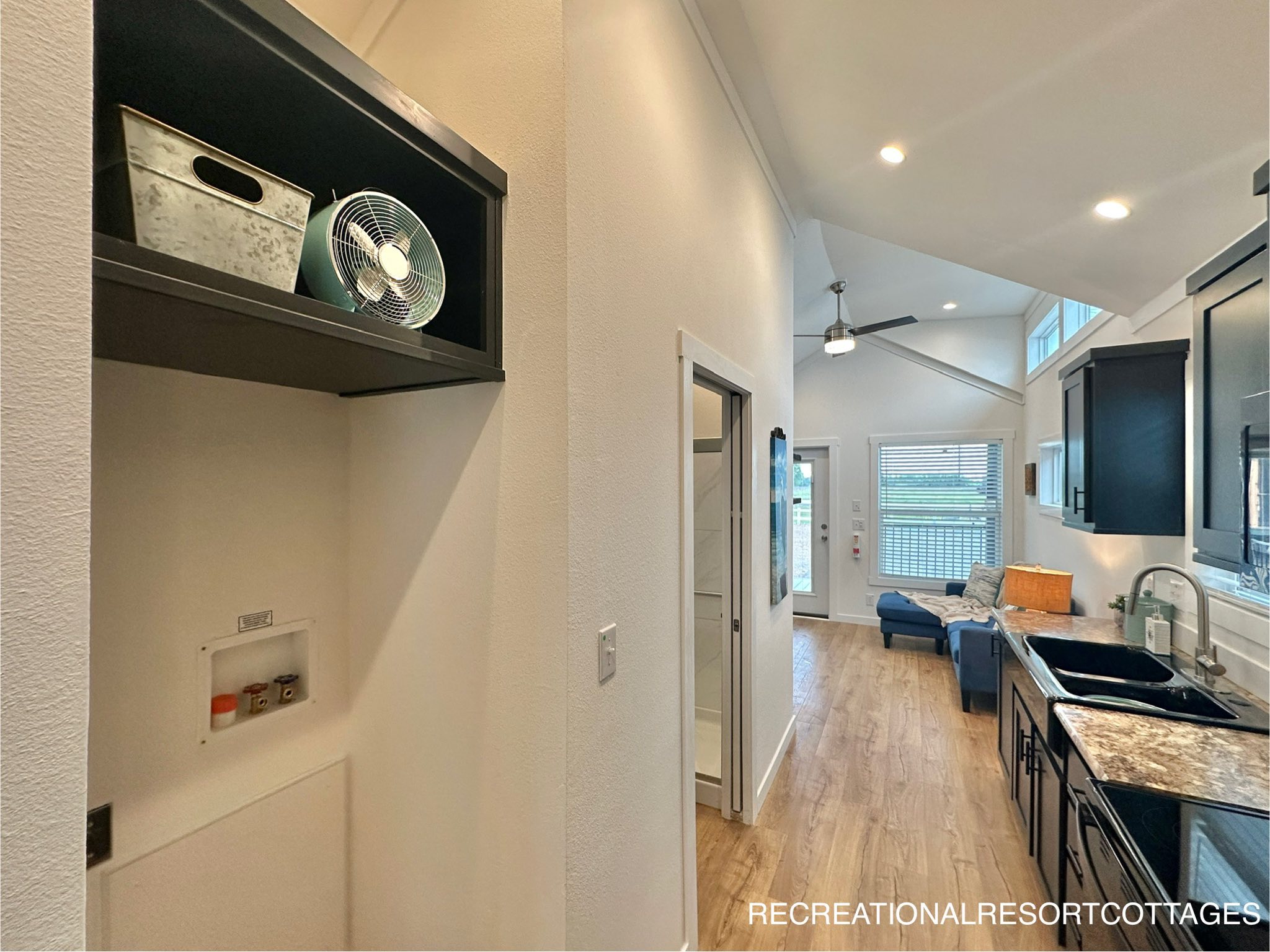 Bright, modern tiny home interior featuring a hallway leading to a cozy living area with large windows. Kitchen has dark cabinets and a double sink.
