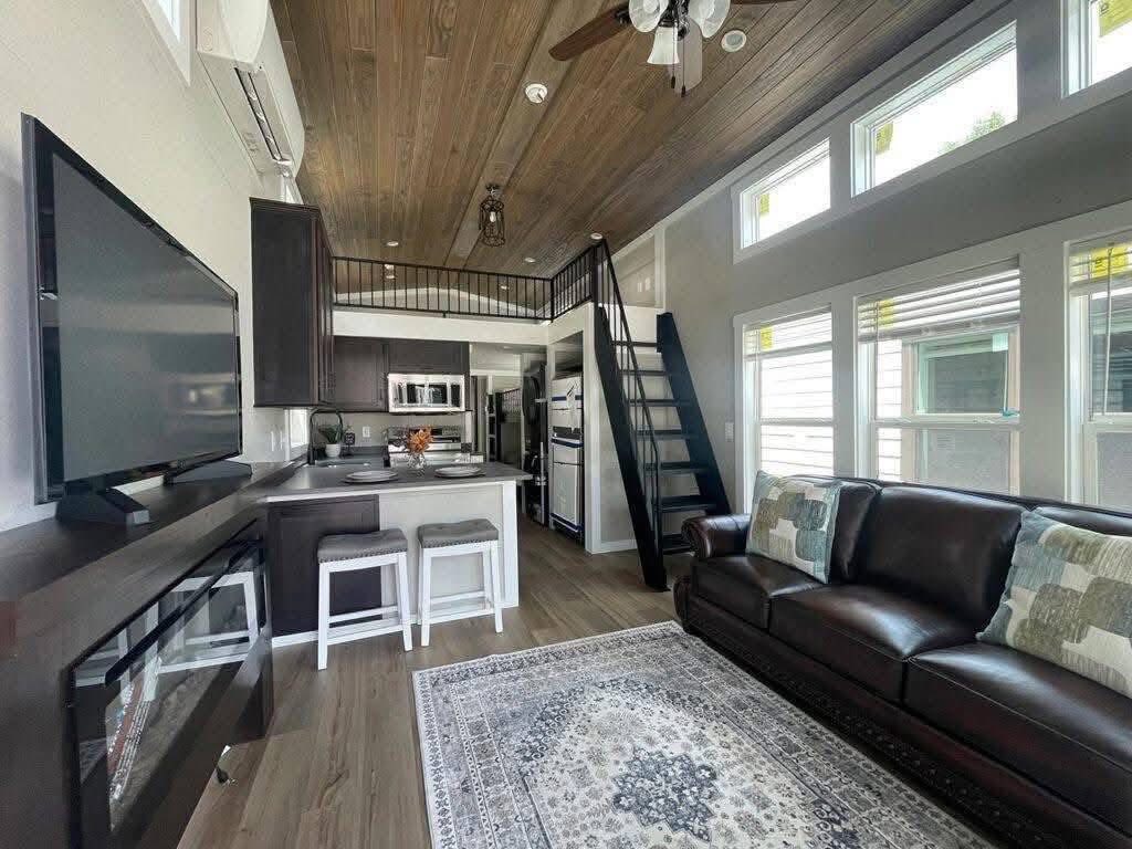 Modern tiny home interior with a cozy atmosphere. Features dark wood accents, a loft with ladder, large windows, leather sofa, TV, and kitchenette.