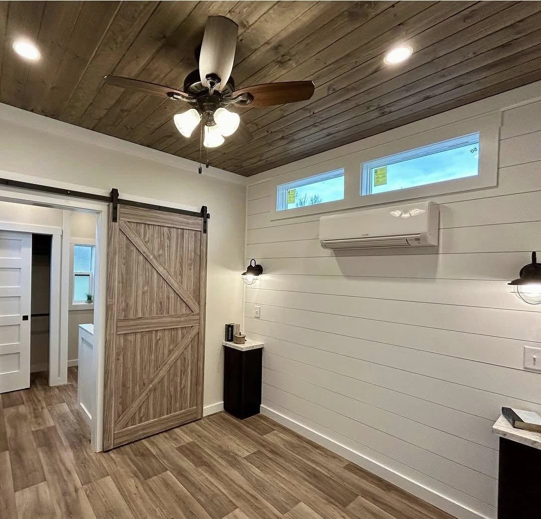 Modern room with wood ceiling and flooring, white shiplap walls, a barn-style sliding door, ceiling fan, recessed lights, and a wall-mounted AC unit. Cozy ambiance.
