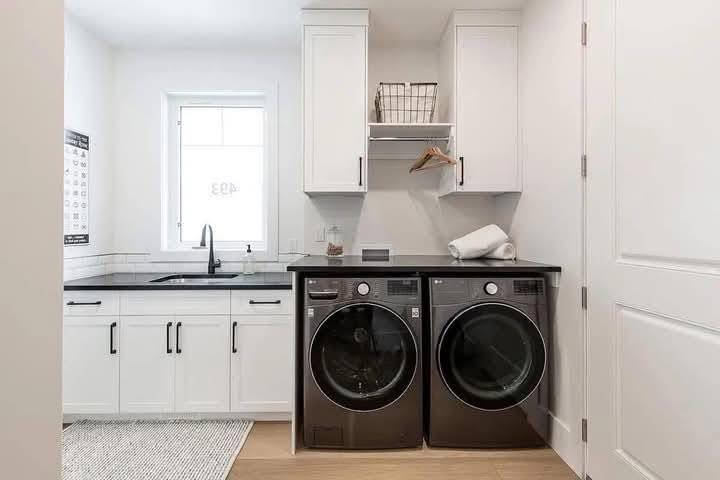 Bright laundry room with white cabinets, black countertop, and a window. Features a washing machine and dryer, shelf with a basket, and folded towels.