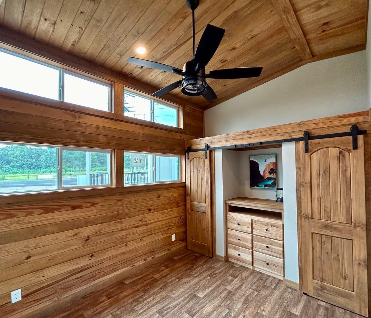 Wooden room with high ceilings, featuring a modern black ceiling fan and large windows on the left. There are sliding barn doors and a built-in dresser with a painting above. The ambiance is cozy and rustic.