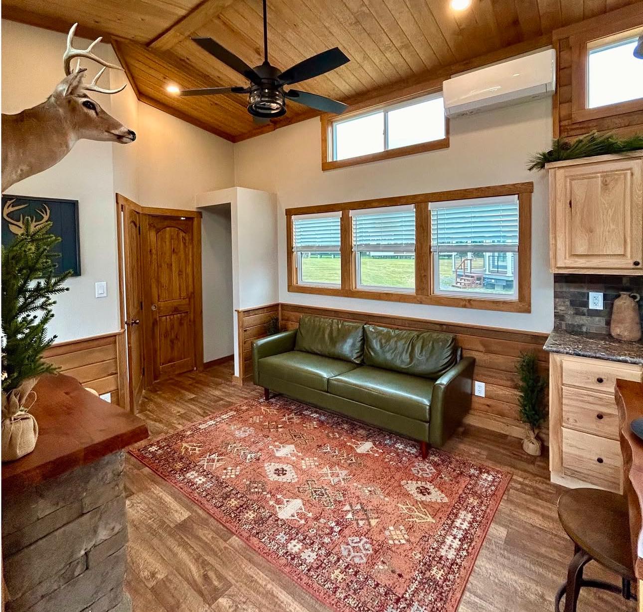 Rustic living room with a green leather sofa, red patterned rug, wooden walls, ceiling, and door. A mounted deer head and a ceiling fan add a cozy, cabin feel.