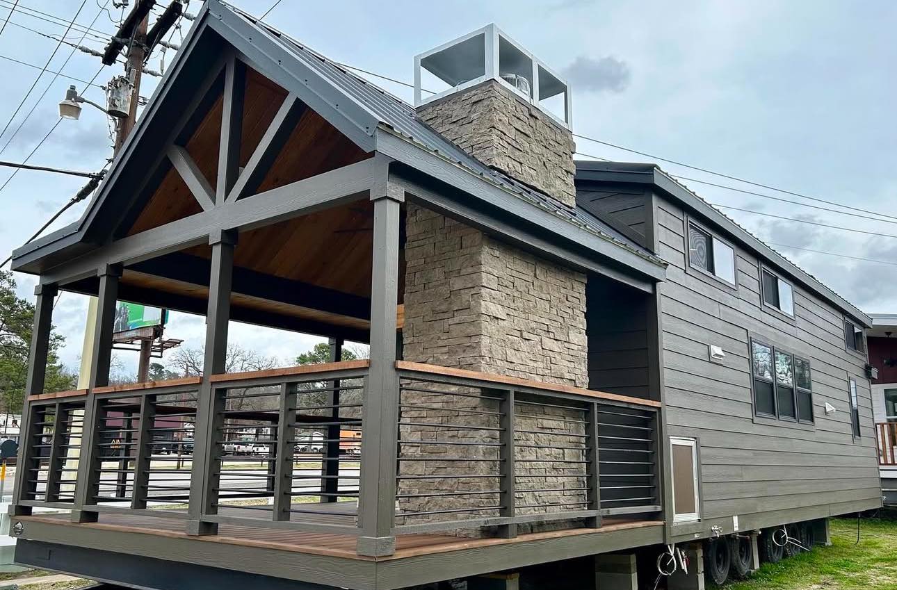 Modern tiny house with gray siding, a stone chimney, and a covered porch featuring wood and metal railings. The overcast sky adds a cozy feel.