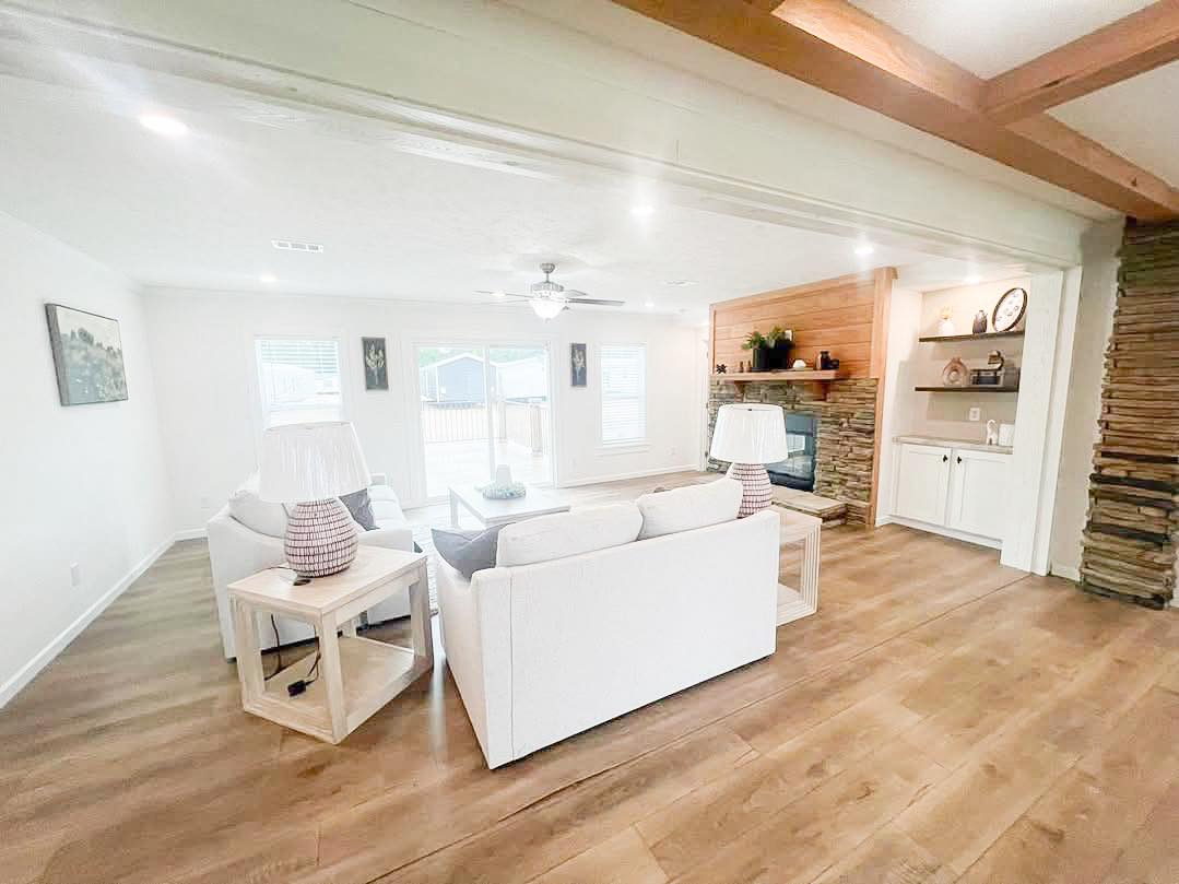 Bright living room with white sofas, wooden floors, and a stone fireplace. Natural light streams through large windows, creating a cozy, inviting atmosphere.