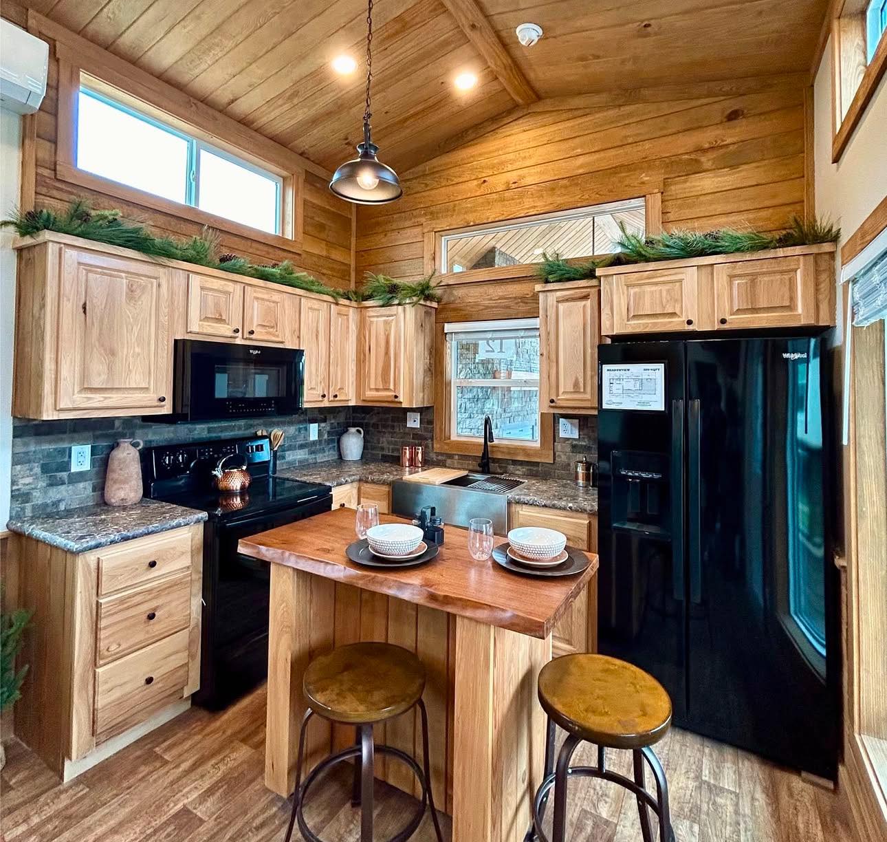 Cozy wood-paneled kitchen with a high ceiling, black appliances, and a center island with stools. Greenery on cabinets adds a rustic touch.