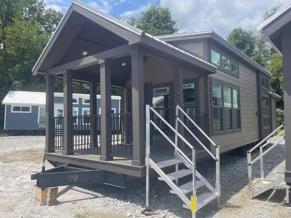 A modern tiny house with a front porch, gray siding, large windows, and metal railings. It sits on a gravel area surrounded by trees, under a partly cloudy sky.