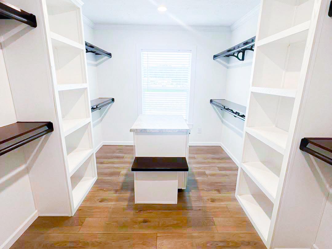 Bright walk-in closet with wooden floors, white shelves, and dark rails lining the walls. A small island with a bench sits at the center.