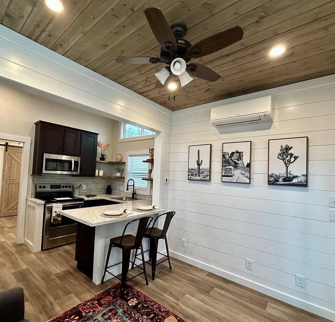 Cozy kitchen with wooden ceiling, white shiplap walls, and a ceiling fan. Features include dark cabinets, a marble island with seating, and nature-themed art.