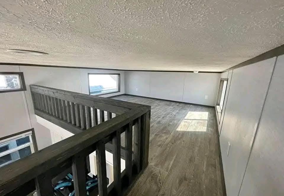 A cozy wooden loft with light gray walls, a low ceiling, and natural light from rectangular windows. The dark wood railing overlooks an open space below.