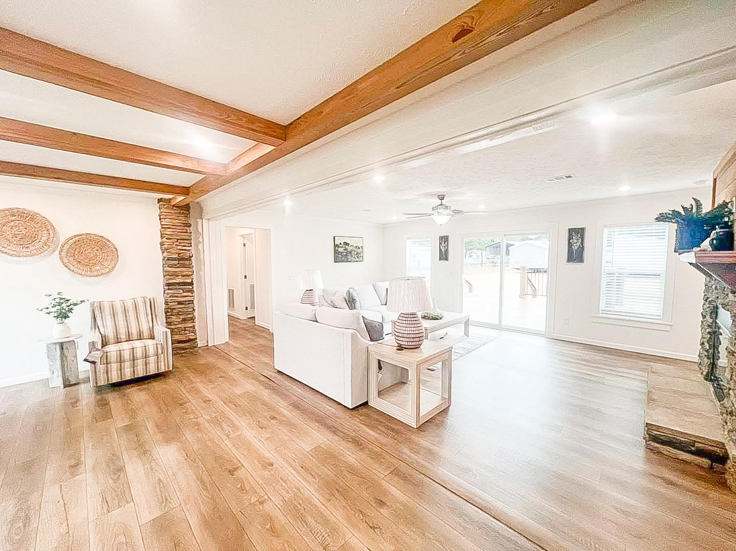 Bright living room with wooden floors, exposed beams, and a stone accent wall. Features a white sofa, striped chair, and large windows. Cozy and airy.