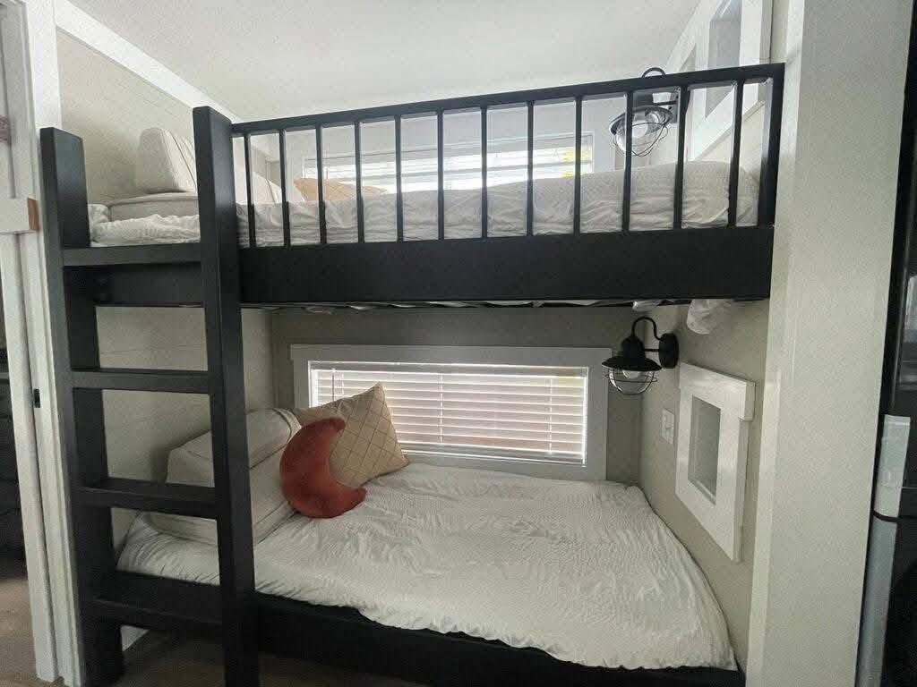 Modern bunk beds in a compact room with white bedding. A ladder leads to the top bunk. A window with blinds and two pillows enhance coziness.