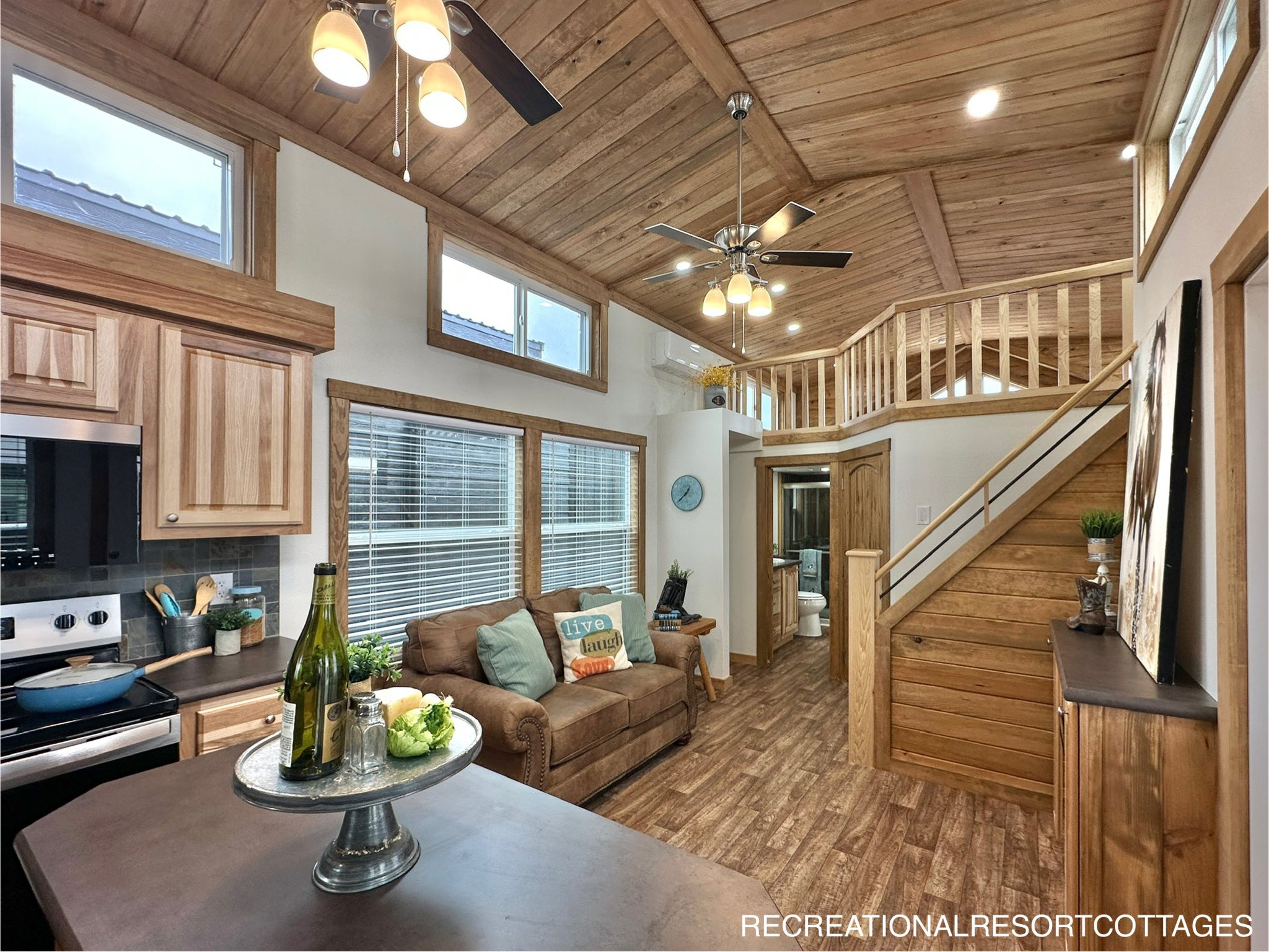 Warm-toned tiny house interior with wooden ceiling and floors. Cozy living area with a brown sofa, modern kitchen, and a loft accessible by stairs.