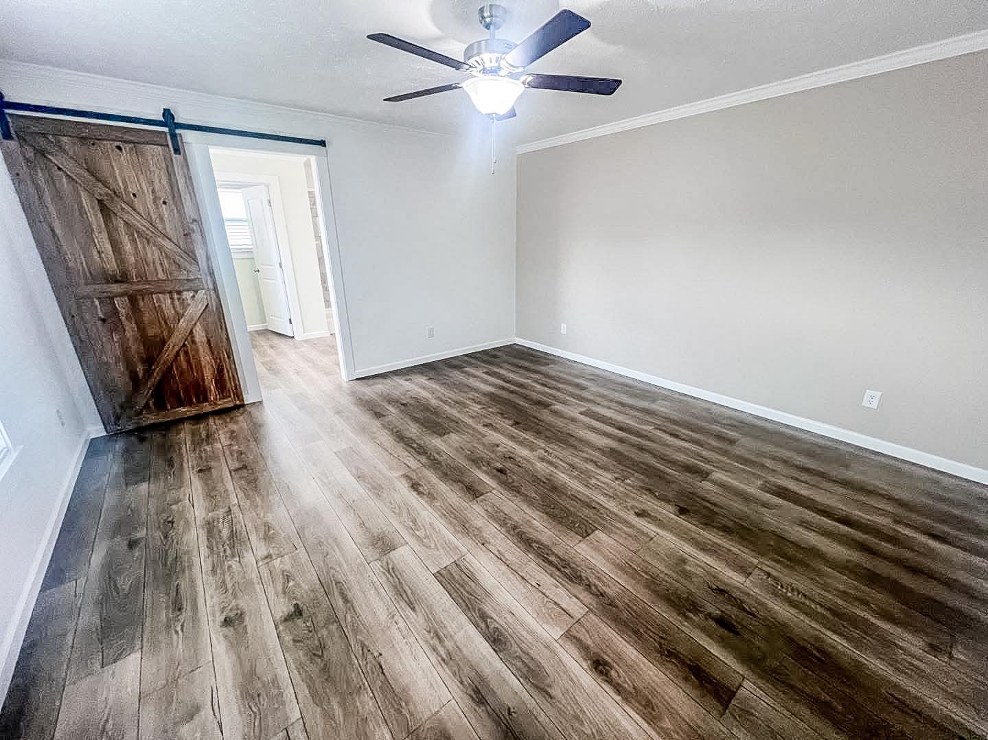A spacious, empty room with light brown hardwood floors and beige walls. A rustic wooden barn door on the left opens to a lit hallway. Ceiling fan overhead.
