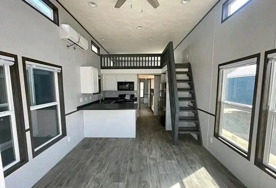 Modern tiny house interior with tall ceilings, large windows, and wood floors. Features a loft accessed by a staircase, and a compact kitchen.