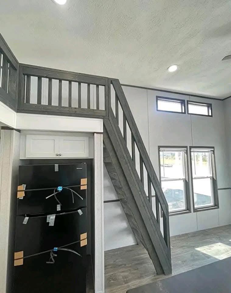 Modern interior with gray stairs leading to a loft. A black fridge is below white cabinets. Large windows illuminate the space with natural light.